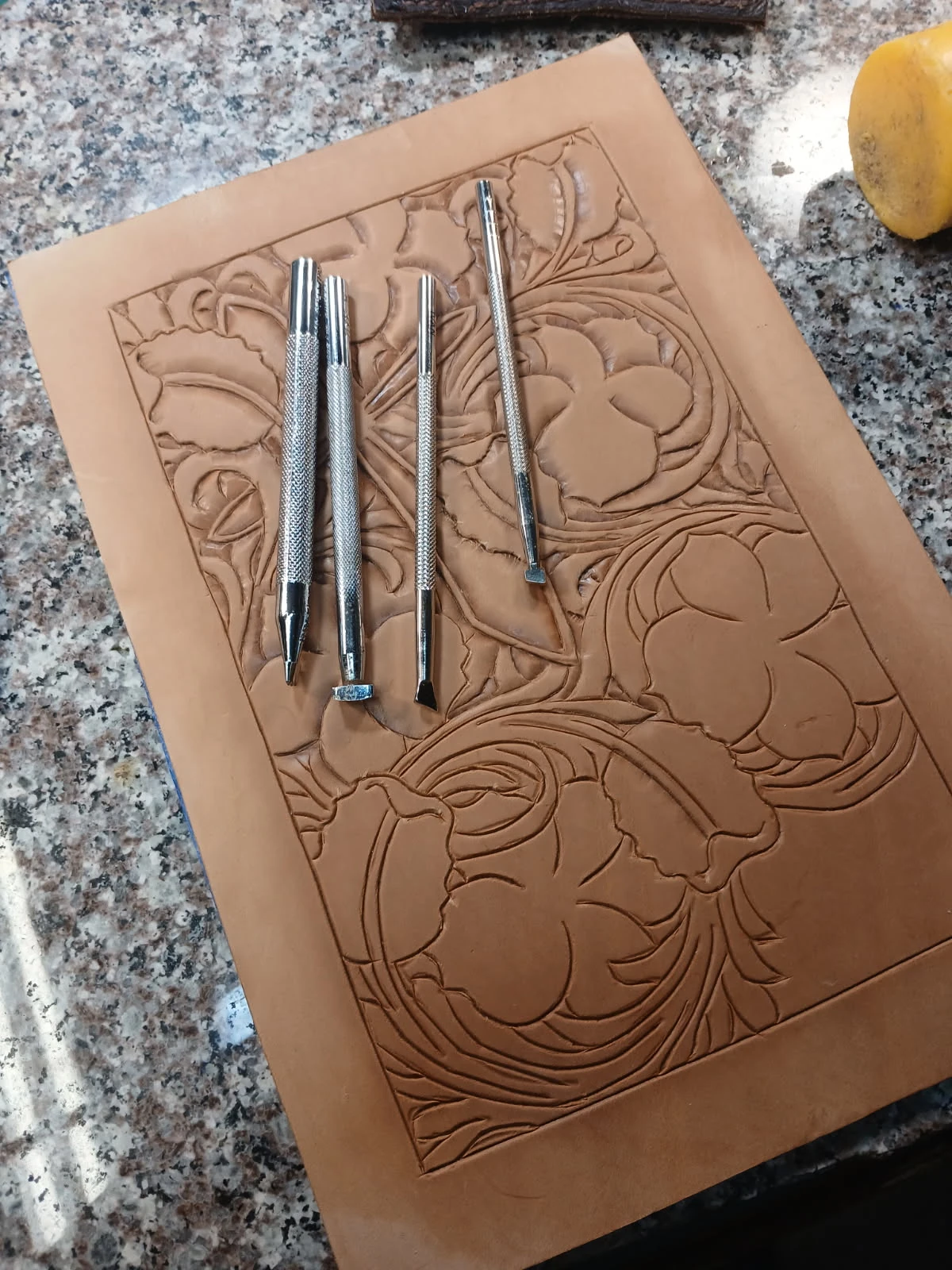 A panel of natural leather with hand-carved Sheridan-style florals, resting on a workbench with four chrome tooling stamps laid across it